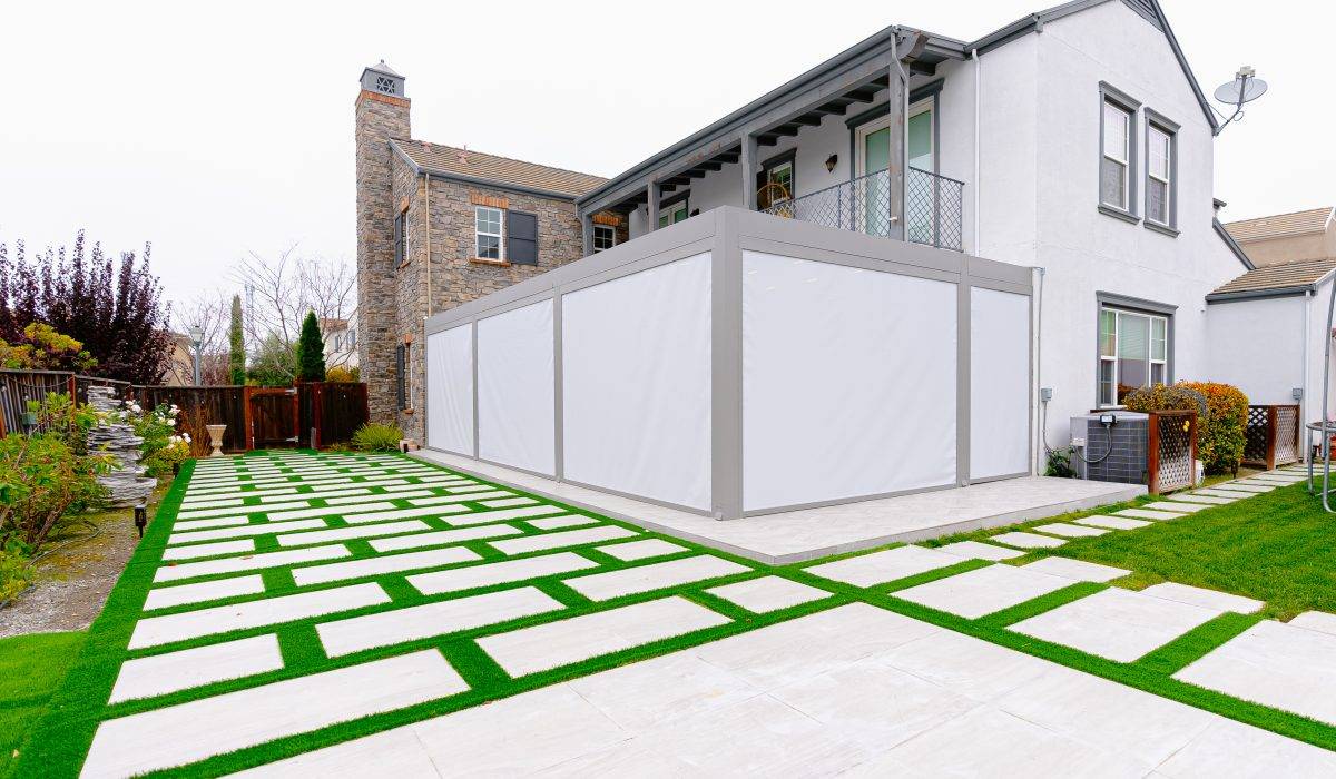 Motorized pergola side enclosure with white screen panels on sleek gray frame beside stone-faced house and grid lawn.