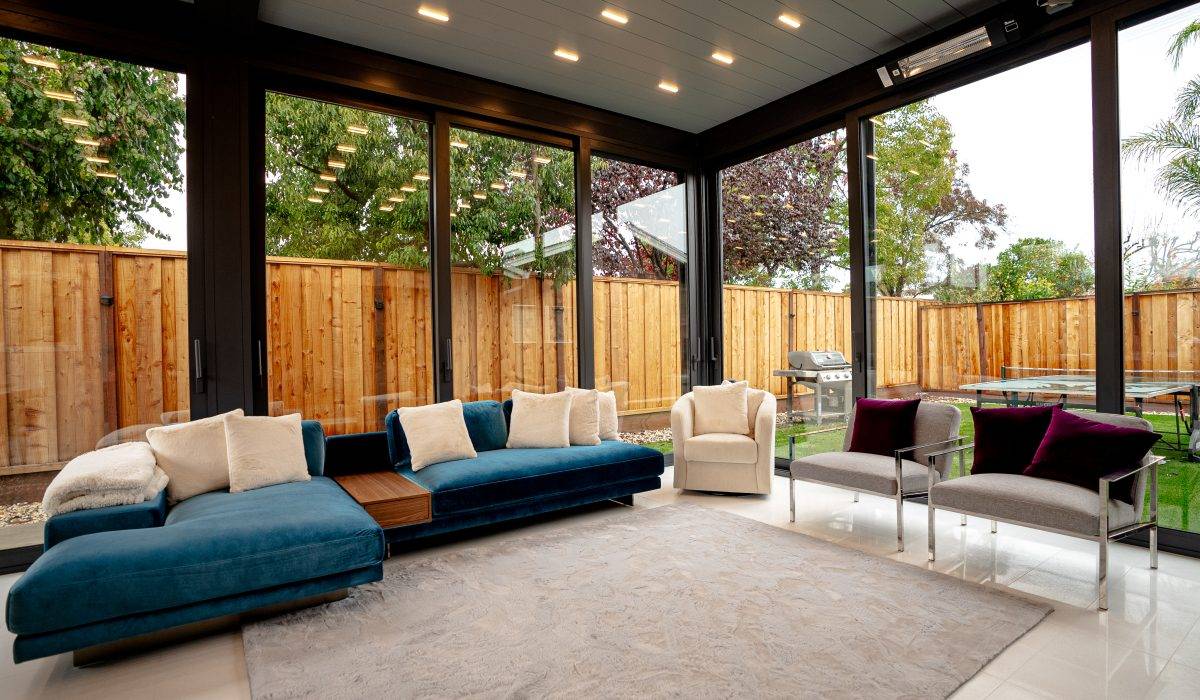 Enclosed pergola lounge with floor-to-ceiling glass panels, ceiling heaters, and blue sectional for year-round comfort.