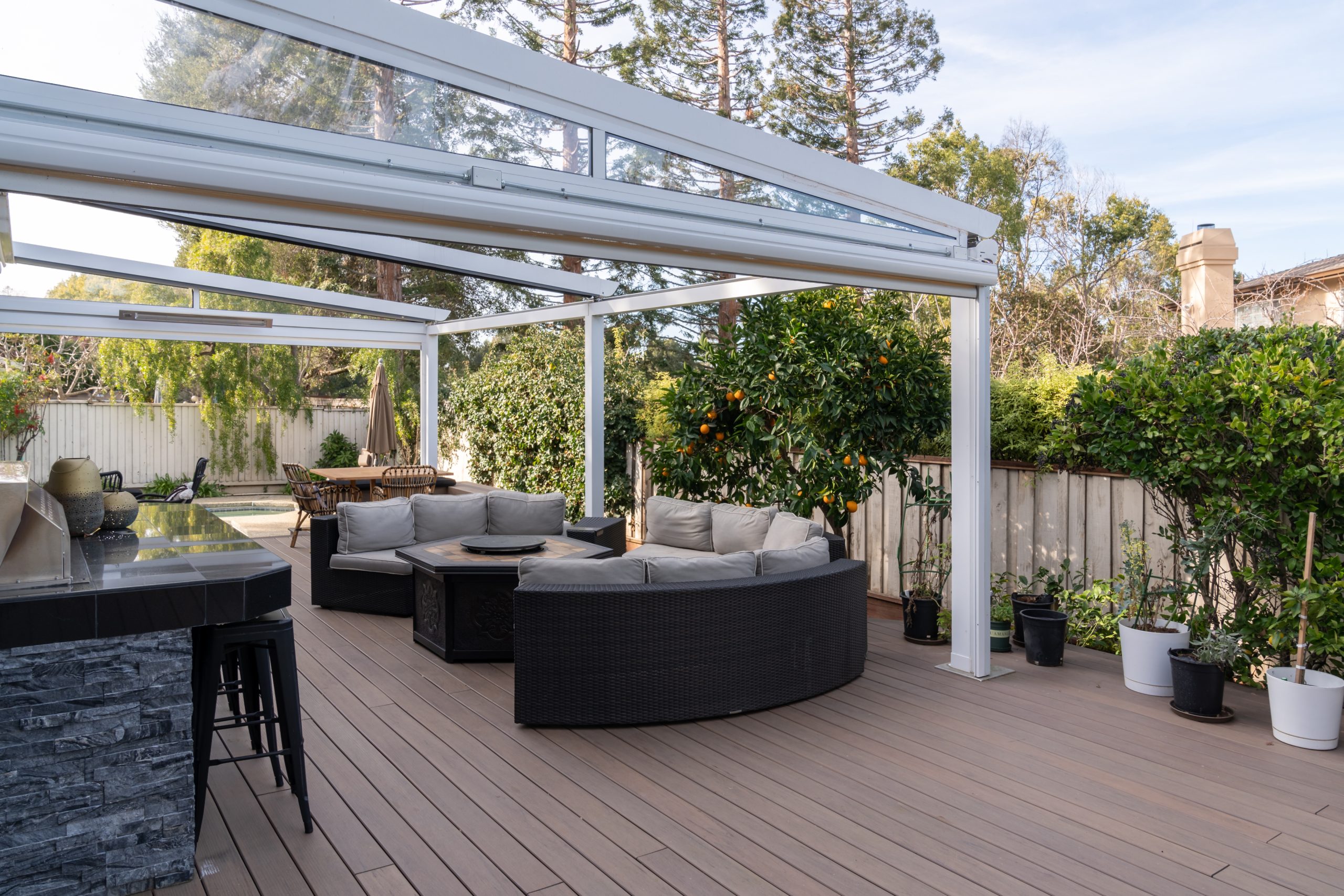 White Pergomatic pergola with slim glass roof panels shades curved rattan sofa and outdoor kitchen on warm wood deck.