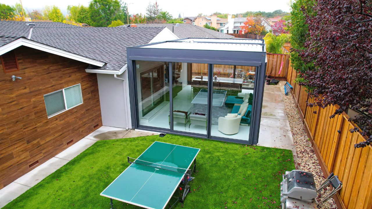 Glass-walled gray pergola room links wood-clad home to yard, sliding panels reveal lounge above green lawn.