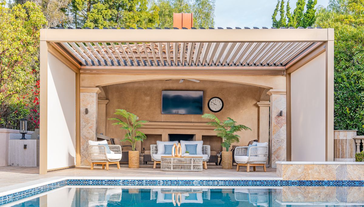 Poolside cabana with beige Louvera pergola with tiltable blades and drop-down screens shelter a TV lounge and wicker seating.