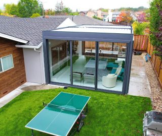 Glass-walled gray pergola room links wood-clad home to yard, sliding panels reveal lounge above green lawn.