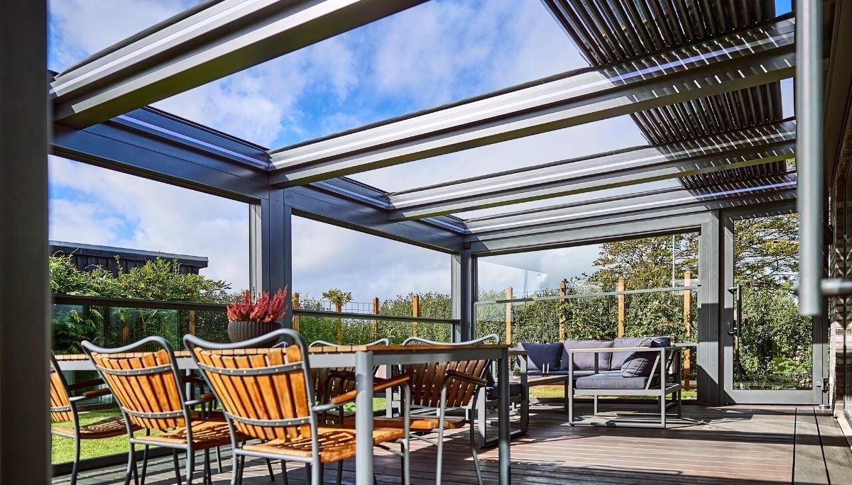 Louvera Glass pergola with retractable glass blades spans modern deck, revealing blue sky above wood-slat dining chairs.